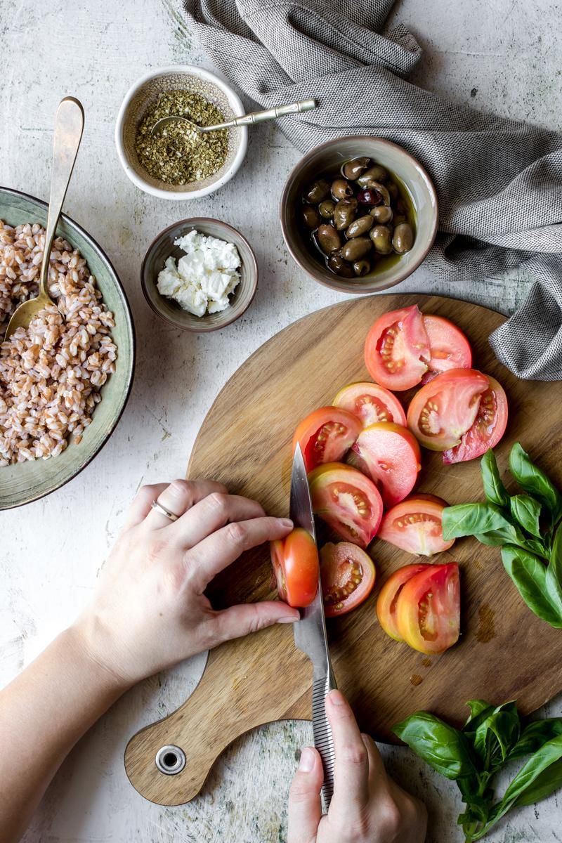 tagliere con due mani che tagliano dei pomodori e basilico, ciotola grande con farro cotto, due ciotole picocle con olive taggiasche e feta sbriciolata