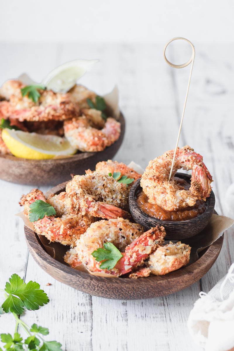 Due ciotole di legno con dentro gamberi impanati low carb al forno e piccola ciotola con finta salsa barbecue, foglie di prezzemolo e fettine di limone e lime, un bastoncino di legno infilzato in un gambero