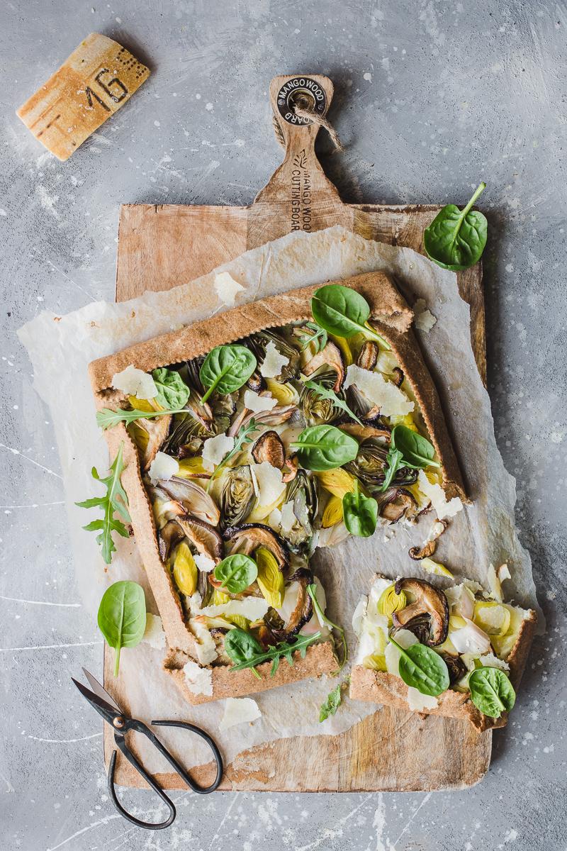 Torta salata integrale senza burro ai porri, funghi shitake e carciofini, con foglie di spinacino e rucola su un tagliere con un paio di forbici e una fetta già tagliata