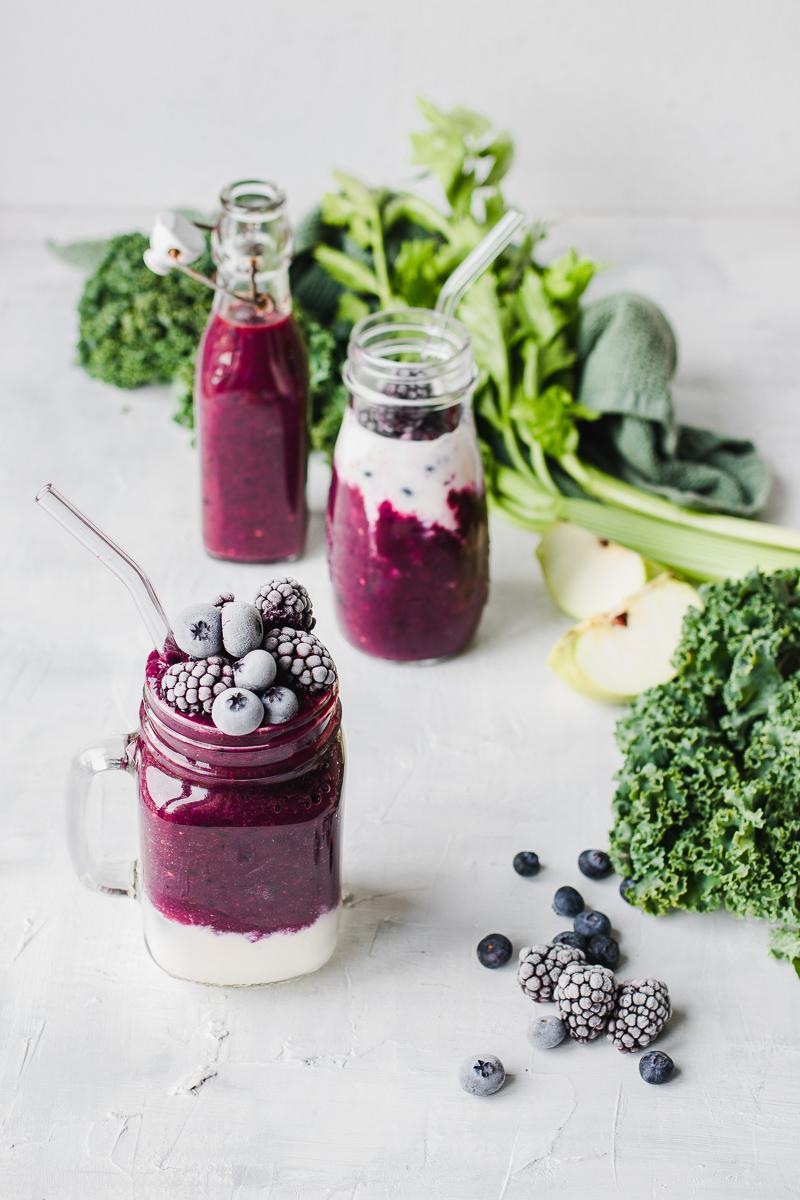 Tre bicchieri di vetro con dentro un frullato viola di verdura e frutta variegato allo yogurt, con foglie di verdura sparse sul tavolo