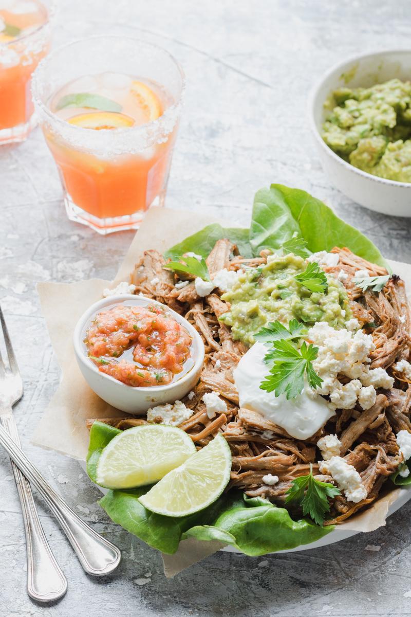 Piatto di carnitas con salsa, ciotola di guacamole e due bicchieri di margaritas in controluce