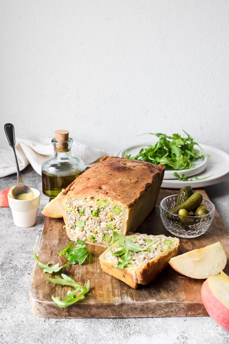 Terrina di maiale in crosta, con pistacchi e piselli, già affettata su un tagliare di legno con mele, sottaceti, rucola e senape