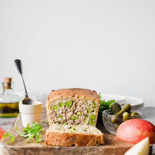 Terrina di maiale in crosta, con pistacchi e piselli, già affettata su un tagliare di legno con mele, sottaceti, rucola e senape