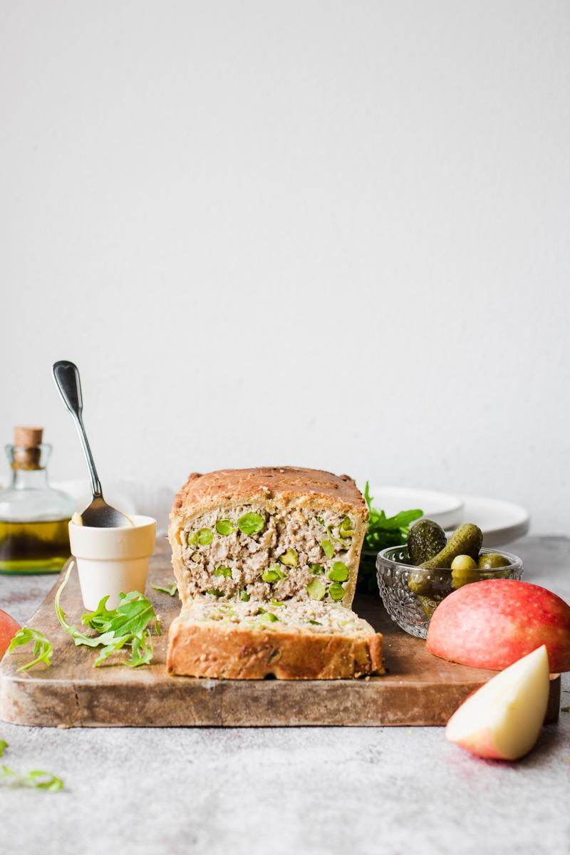 Terrina di maiale in crosta, con pistacchi e piselli, già affettata su un tagliare di legno con mele, sottaceti, rucola e senape