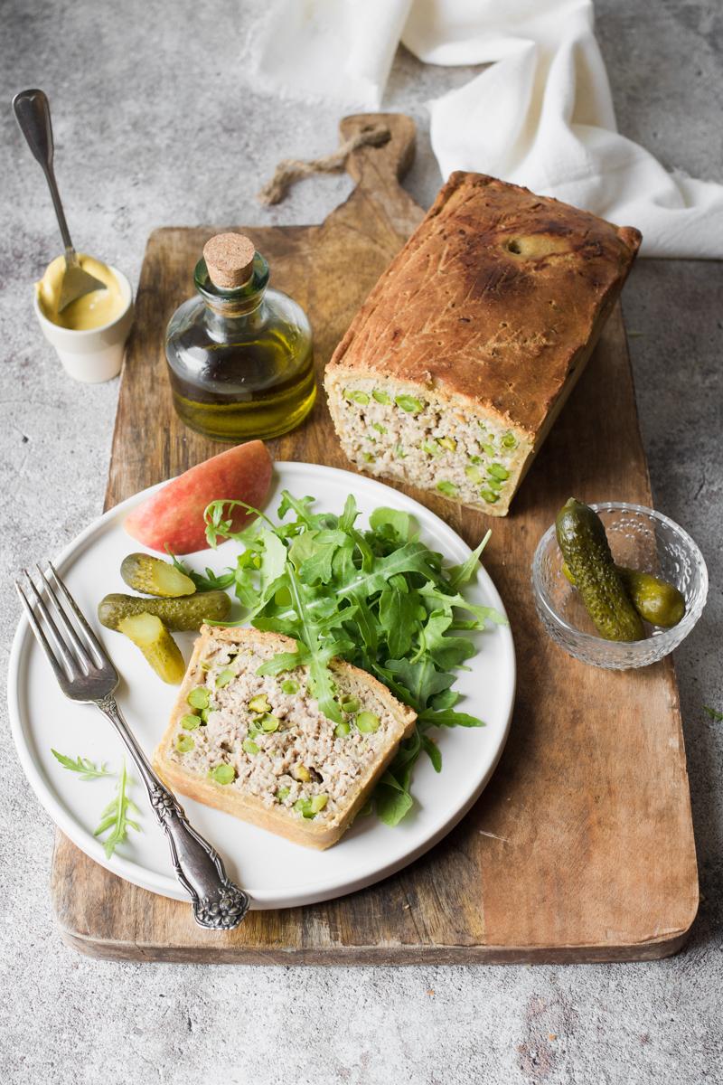 Terrina di maiale in crosta, con pistacchi e piselli, già affettata su un tagliare di legno con mele, sottaceti, rucola e senape