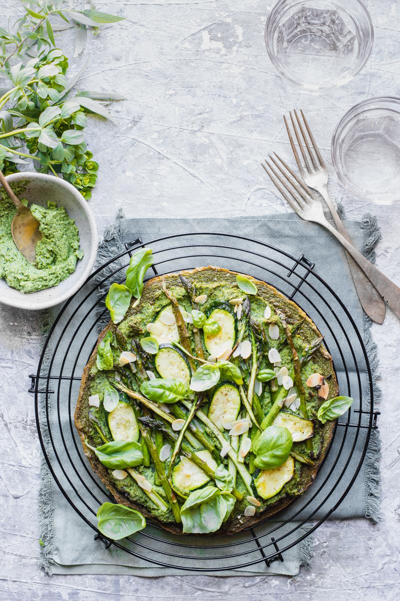 Socca verde rotonda con verdure su una grata e ciotola di pesto di broccoli accanto, con forchette