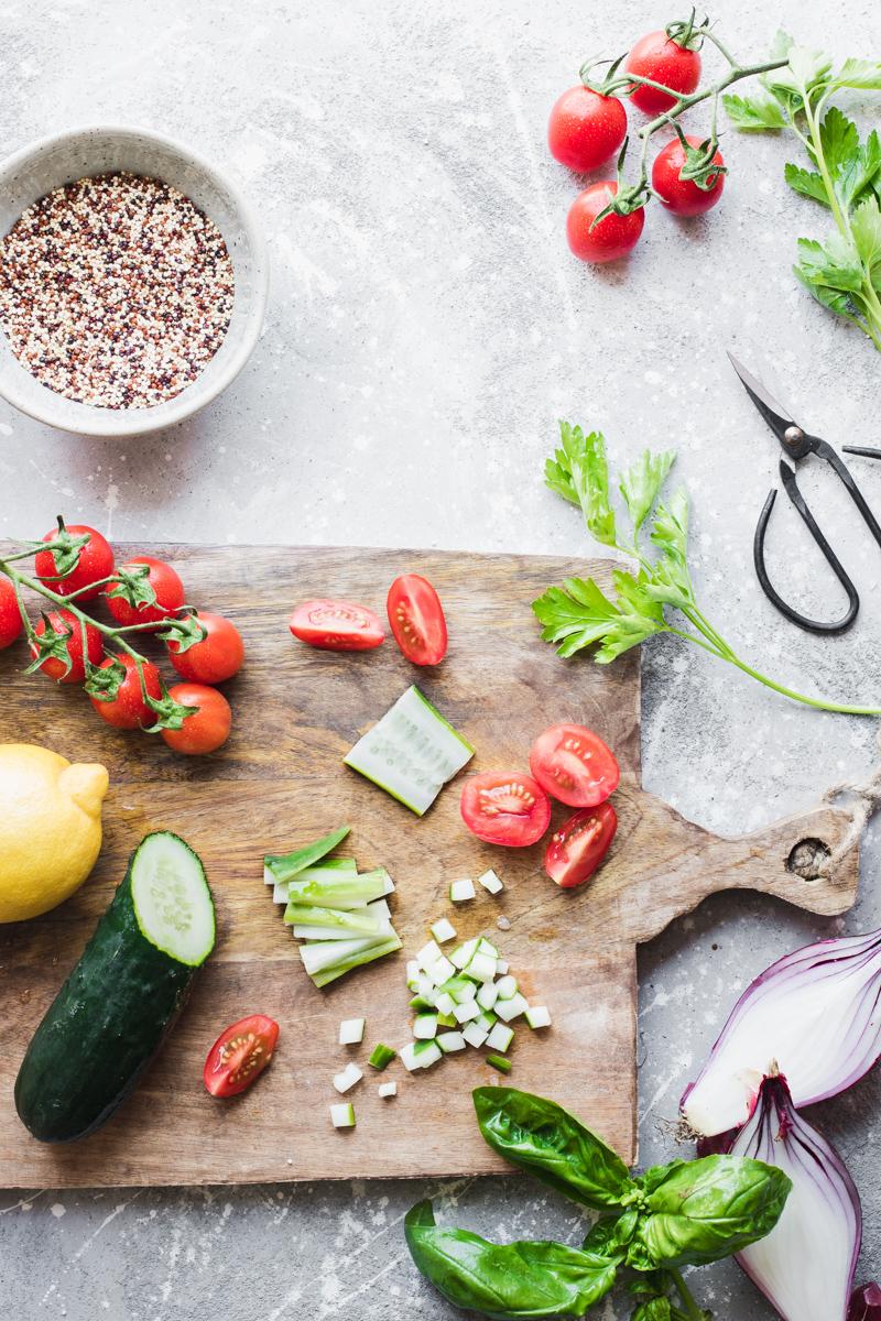 Tagliere di legno con pomodori, cetrioli, limone, erbe aromatiche, cipolla rossa e ciotolina con quinoa tricolore cruda