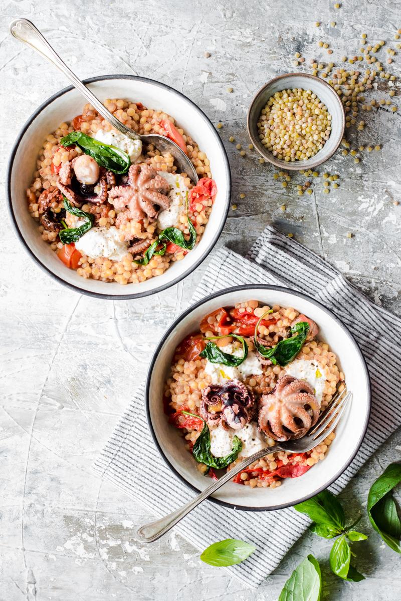 Due ciotole piene di fregola sarda, moscardini, pomodori, burrata e basilico, con uno strofinaccio a righe sotto e foglie di basilico