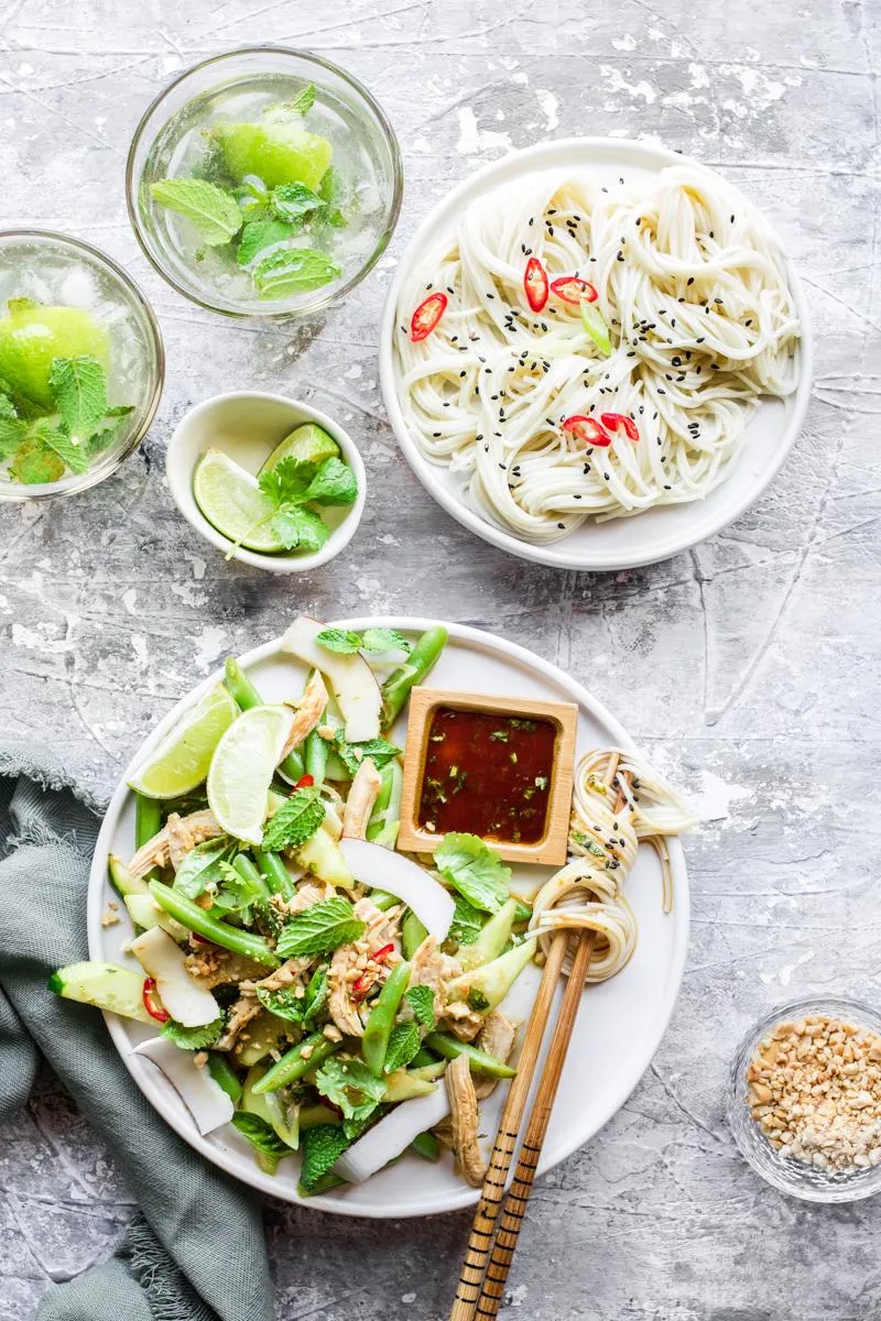 Piatto con verdure verdi, insalata di pollo, bacchette con noodle avvolti e piattino quadrato di salsa di soia, accanto a un piatto di noodle e un bicchiere di mojito analcolico