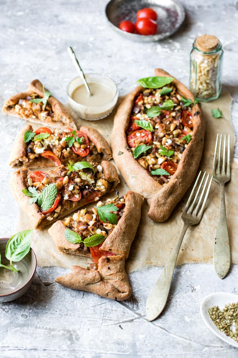 Foglio di carta da forno con due pizze turche con pomodorini, ciotolina con origano secco e posate, vista a 45°