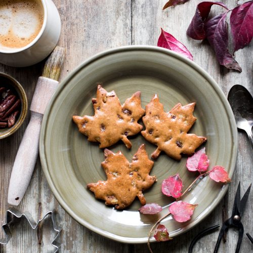 tre biscotti di farina di castagne e sciroppo d'acero senza glutine a forma di foglia su un piatto visto dall'alto con attorno foglie autunnali e un caffè