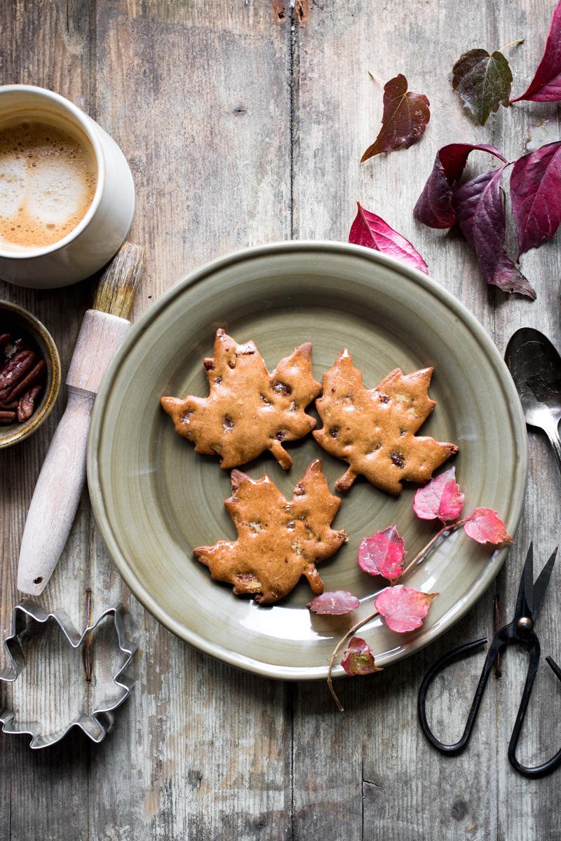 tre biscotti di farina di castagne e sciroppo d'acero senza glutine a forma di foglia su un piatto visto dall'alto con attorno foglie autunnali e un caffè