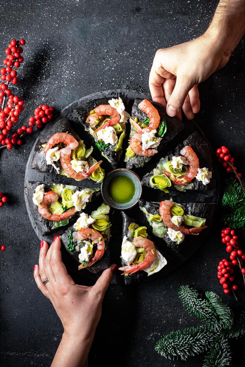 Piatto di ardesia con spicchi di focaccia al nero di seppia con gamberi rossi crudi e burrata, due mani che prendono due fette