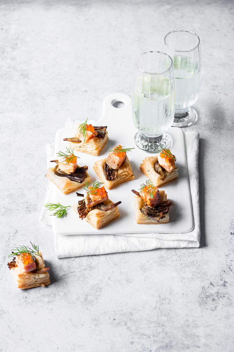 Salatini di sfoglia con salmone e carciofi su un vassoio di ceramica bianca con tovagliolo e due bicchieri