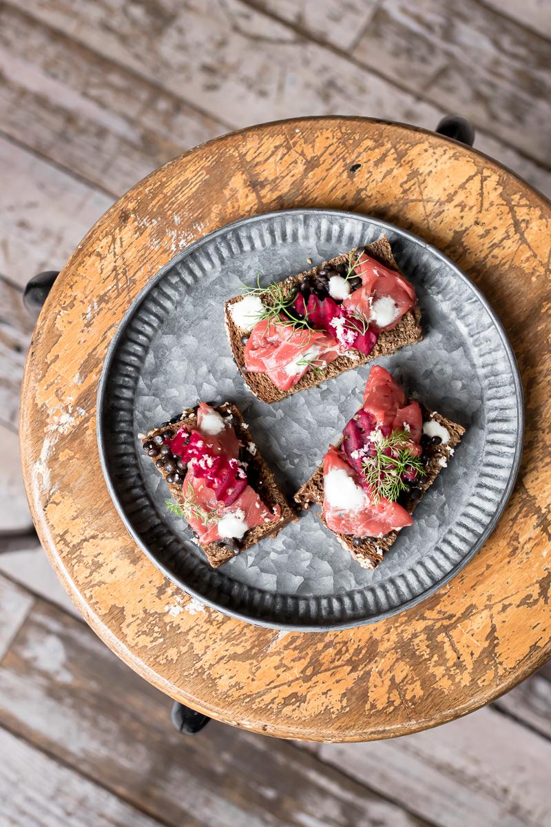 Piatto zincato con tre tartine di pane nero con carpaccio, lenticchie e barbabietola, su uno sgabello di legno