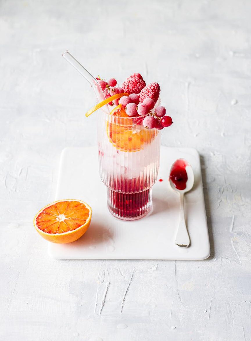 Vassoio in ceramica bianco con bicchiere da cocktail con gin tonic al melograno e araancio rosso, ribes congelati, cucchiaio bianco sporco di melograno e mezza arancia