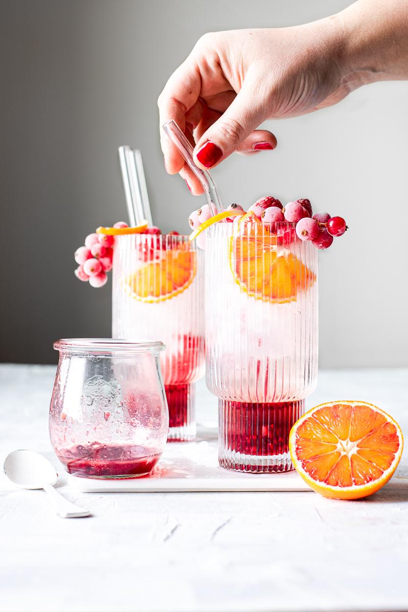 Mano che prende una cannuccia di vetro immersa in un bicchiere di kir gin tonic con frutti di bosco congelati