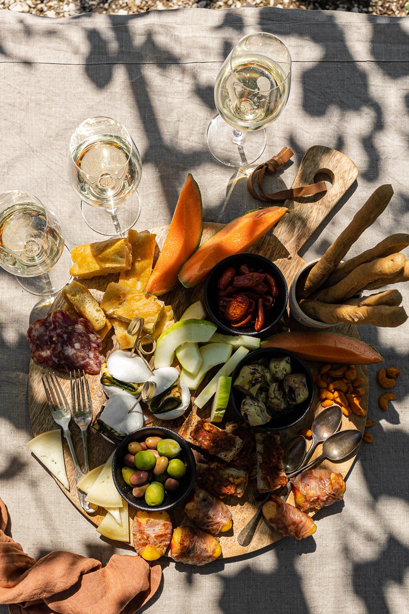 tagliere da aperitivo estivo pieno di cibo su tovaglia rustica con bicchieri di vino e ombre di viti